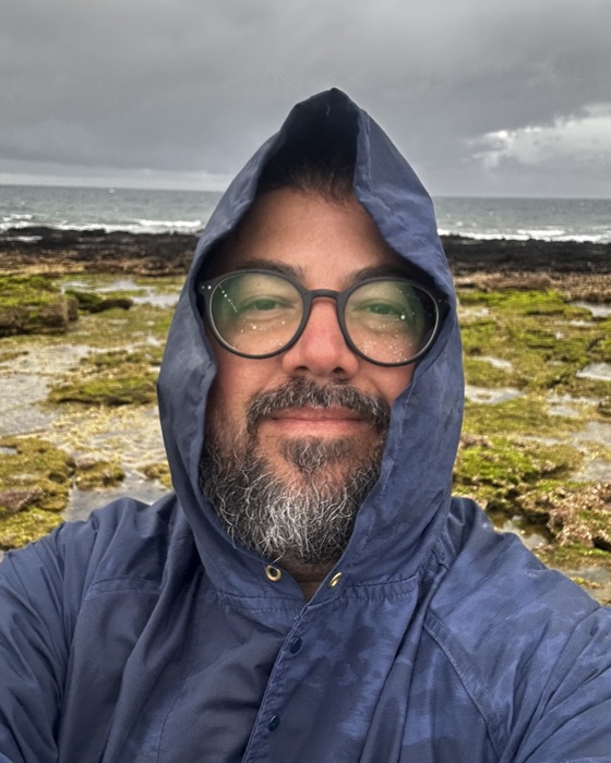 Juan Paz on a rocky shore, wearing a hooded jacket, on an overcast day